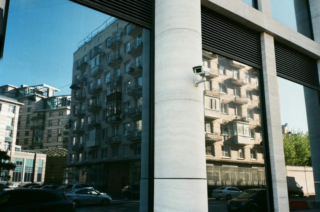 Tall beige multi storey dwelling house reflecting in window of modern construction with small security camera mounted on thick white column